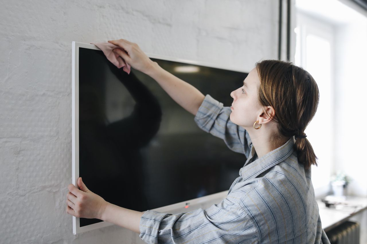 Woman wiping flat screen TV indoors, focusing on cleaning and maintenance.